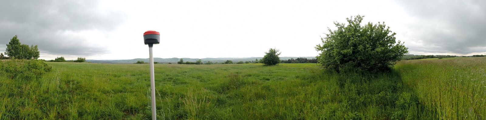 Panorama rozległej łąki z wysoką trawą; na pierwszym planie odbiornik GNSS na tyczce pomiarowej, w tle pas drzew i pagórki pod zachmurzonym niebem